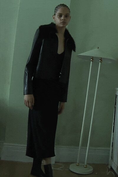 Black leather jacket with a broad furry collar and glossy sleeves. Black scoop-neck column dress falling straight to the lower calf. Matte black ankle boots with a close, socklike shaft. Standing in a rented flat before dawn, radiator ticking against the wall.