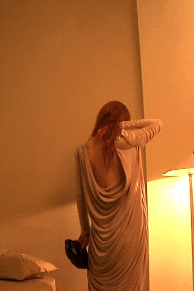 Blush jersey gown with a plunged open back and pooled cowl drape. Black beaded clutch tucked low in one hand. Hotel lamp glow, halfway through taking your earrings off.