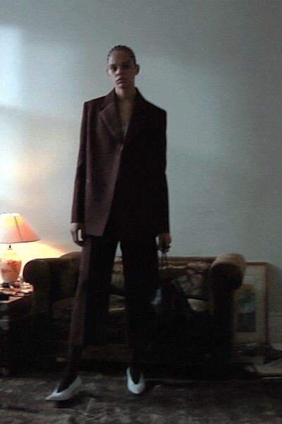 Oxblood blazer with hard peaked lapels and a long single-breasted line. Matching straight trousers falling clean over the shoe. Black carryall with short handles hanging from one hand. White pointed pumps catching the light at the vamp. Whatever you wear back to the hotel after missing the last dinner course.
