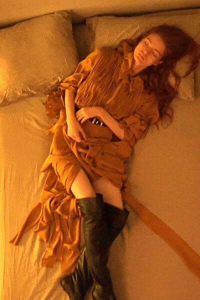 Ochre draped dress with a ruched bodice and ruffles collapsing at the cuffs. Wide self belt knotted low, one long tail slipping off the bed. Black knee-high leather boots with soft shafts and blunt toes. Morning after a hotel wedding, curtains still shut against the sun.