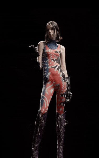 Skin-tight printed unitard with a high blue mock neck. Black leather gloves, one elbow-length and one cut shorter. Purple knee boots with pale whipstitch lacing climbing the front. Under a strip-mall sign after midnight, still too wired to go home.