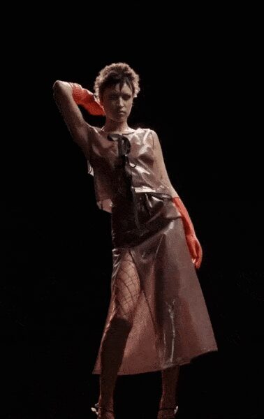 Opera-length orange gloves in glossy rubber shine. Silver metallic sleeveless top, tied at the chest with black lacing. Shiny black apron panel over a translucent midi, fishnet leg showing through. Thin black ankle-strap sandals. Whatever survives the walk home after the club lights snap on.
