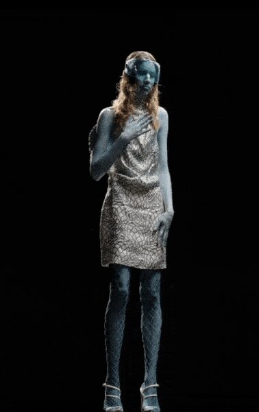 Silver sleeveless shift in crackled metallic jacquard, draped softly at the neck. Pale lace-pattern tights. Silver ankle-strap heels. Under stage haze, as if frost climbed up from the floor.