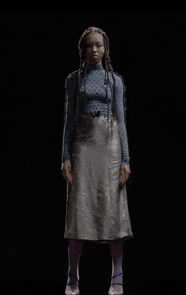 Powder-blue fitted knit top with a high neck and long sleeves. Silver column skirt in crushed metallic fabric, falling to mid-calf. Muted violet opaque tights. Silver ankle-strap heels with a low shine. Early train platform, frost in the air and nowhere warm yet.