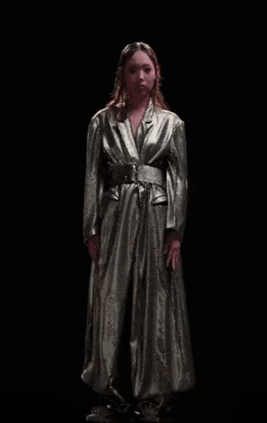 Liquid silver lamé jumpsuit with a revere collar and pooled wide legs. Wide dark belt cinching hard, with flap pockets jutting at the hips. Silver shoes barely visible under the dragging hem. Under the gym stage lights after the chairs have all been stacked.