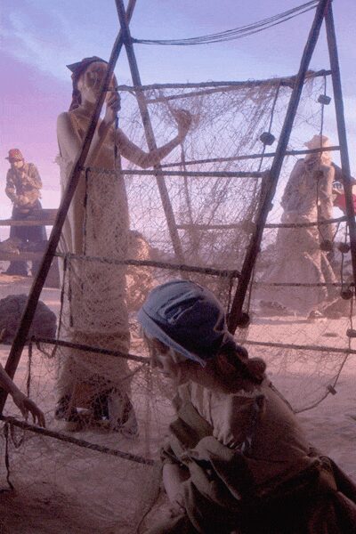 Long bone lace dress, column-cut with a small flutter at the sleeve. Dark narrow belt cinching the waist. Powder-blue scarf tied low over the hair, knot at the nape. Caught inside a netted enclosure while the fairground dust keeps lifting.