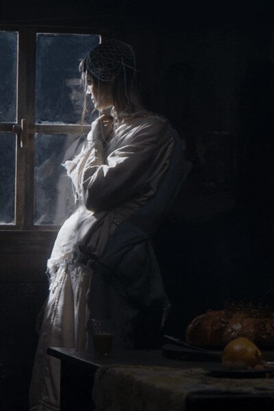 Fine net veil draped over the hair, catching the window light. Ivory satin robe loosely wrapped, frayed ruffles breaking at the cuffs. Long pale skirt falling in a soft column beneath the robe. Cold kitchen before sunrise, bread still on the table from last night.