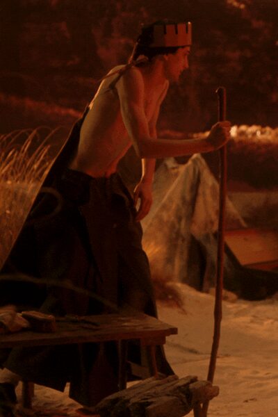 White paper crown with blunt castle points circling the head. Sleeveless ivory knit top, cut close with the armhole sinking low. Dark full skirt pooling heavy at the ground, apron flap hanging loose. Long wooden staff carried upright in one hand. Firelight on packed sand, halfway through a pageant nobody rehearsed enough.