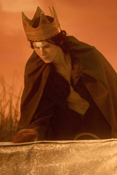 Creased brown paper crown with hand-drawn medallions at each point. Heavy dark cape thrown over both shoulders, one edge folding inward. Open-neck pale shirt slipping loose beneath the drape. Black trousers disappearing into the low light. Kneeling on a banquet table after the pageant has emptied out.