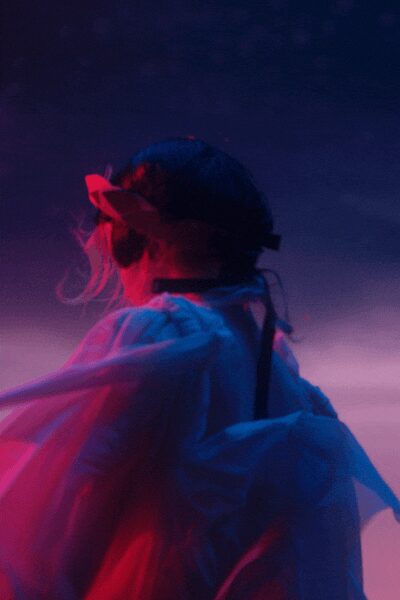 Oversized bow tied over cropped dark hair. Sheer white organza blouse, sleeves ballooning into the colored light. Wide black choker, blunt against the throat. Single black strap cutting straight down the back. At the fair after dark, wind lifting sugar smoke off the stalls.