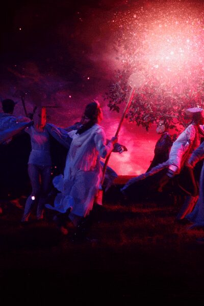 Long white coat flaring open, soft fabric catching blue and magenta light. Pale ruffled dress underneath, hem breaking into loose layered frills. Dark ankle boots partly swallowed by the grass. Running across wet ground under a flare while everyone else scatters.