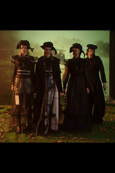 Black felt bonnet with long ties drooping beside the face. Sheer black long-sleeve top traced with pale piping and a narrow white belt. Smoky tulle skirt over checked panels, one white patch catching at the shin. Dark knee boots under the transparent hem. Crossing wet grass after dusk while the house lights burn behind them.