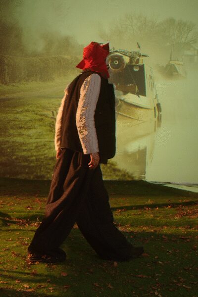 Red pointed hood wrapped close, the tip jutting backward. Pale pinstriped shirt with slim vertical lines and loose cuffs. Long black fleece vest, cut boxy and left open. Dark wide-leg trousers pooling heavily over the shoes. Crossing damp grass beside a canal boat throwing up cold spray.