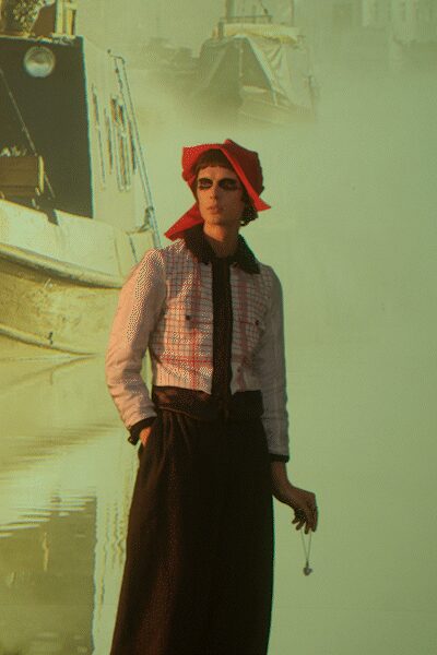Red scarf tied into stiff winged corners around a black bob. Pale pink blouse with a checked bib front and black tie placket. High-waisted black wide-legs falling in a long uninterrupted column. Single drop earring, bead hanging from a fine wire. Harbour fog before noon, standing where the boats come in.
