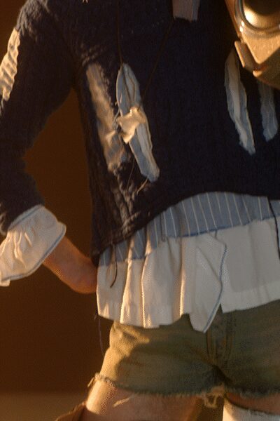 Navy cable-knit sweater with pale ribbon ties dangling at the chest. Blue-and-white striped shirting peeking out in a wide ruffled hem. Olive cutoff shorts with frayed leg openings and a low rise. Walking from the bonfire back to the car with sand in your cuffs.