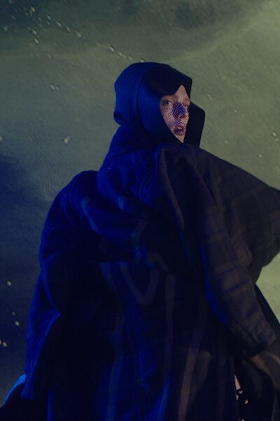 Black hood framing the face close, cut with a deep cowl-like opening. Fine mesh veil over the face, dotted and slightly glittering. Voluminous black cloak with wide checked panels falling in heavy folds. Crossing a floodlit embankment after midnight, wind pushing the cloth sideways.