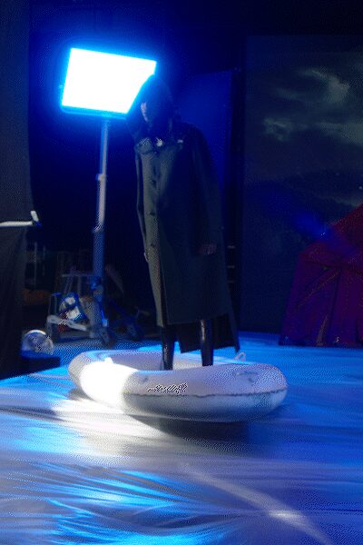 Long black overcoat with a double row of dark buttons. Narrow black trousers disappearing into the light. Black shoes mostly obscured by the inflatable raft. White vinyl dinghy worn around the feet, sagging at one side. Crossing a flooded stage after the generator kicked back on.