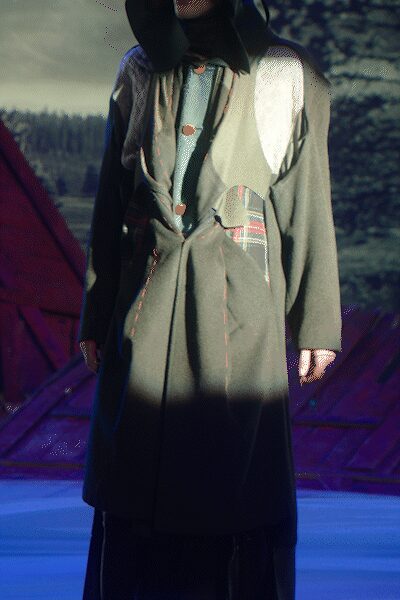 Wide black felt hat shading the face. Moss wool coat with cream shearling bib and plaid patch pocket. Pale blue knit dress peeking through the open front. Dark loose trousers pooling below the hem. Cabin porch after rain, someone still carrying wood in from the trees.