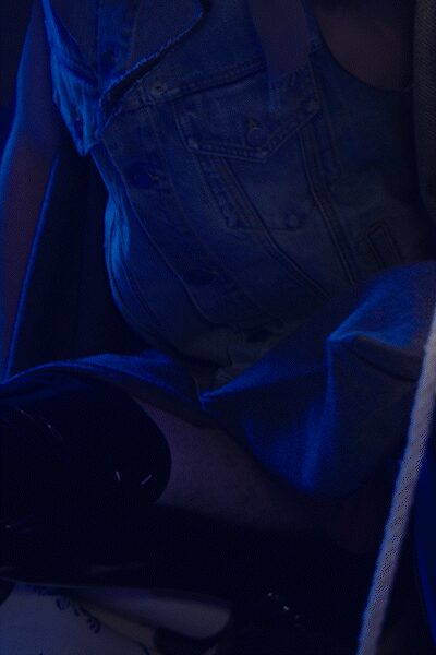 Light-wash denim vest with raw armholes and flap chest pockets. Dark scoop-neck tank barely visible under the open front. Pale denim shorts, soft creases catching the blue club light. Black patent knee-high boots with a hard mirror shine. Sunk into the back booth after midnight, coat sliding off one shoulder.