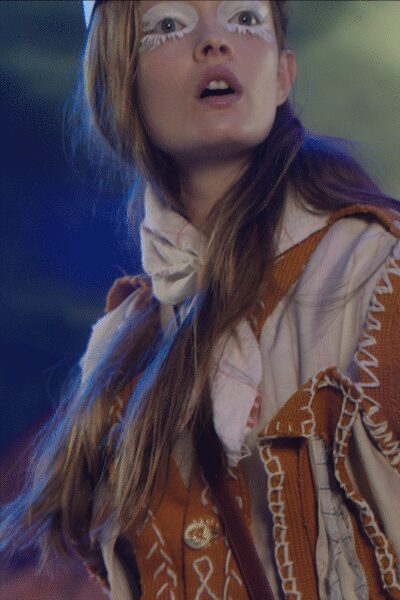 Ivory high-neck blouse with an oversized bow knotted under the chin. Rust suede vest stitched with white whipstitch and small geometric embroidery. Cream patchwork jacket with raw-edged seams and zigzag trim running the sleeve. Bonfire smoke in your hair after the fairground generators cut out.