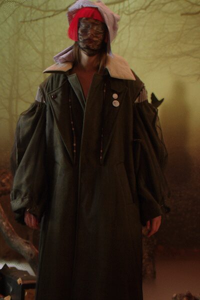 Red knit hood piled with a lavender striped shirt wrapped on top. Brown mottled mesh veiling the face. Long swamp-green coat with dropped cutout sleeves and broad shearling collar. Three white badges pinned high on the chest. Deep in the fog machine woods after the bonfire burned down.