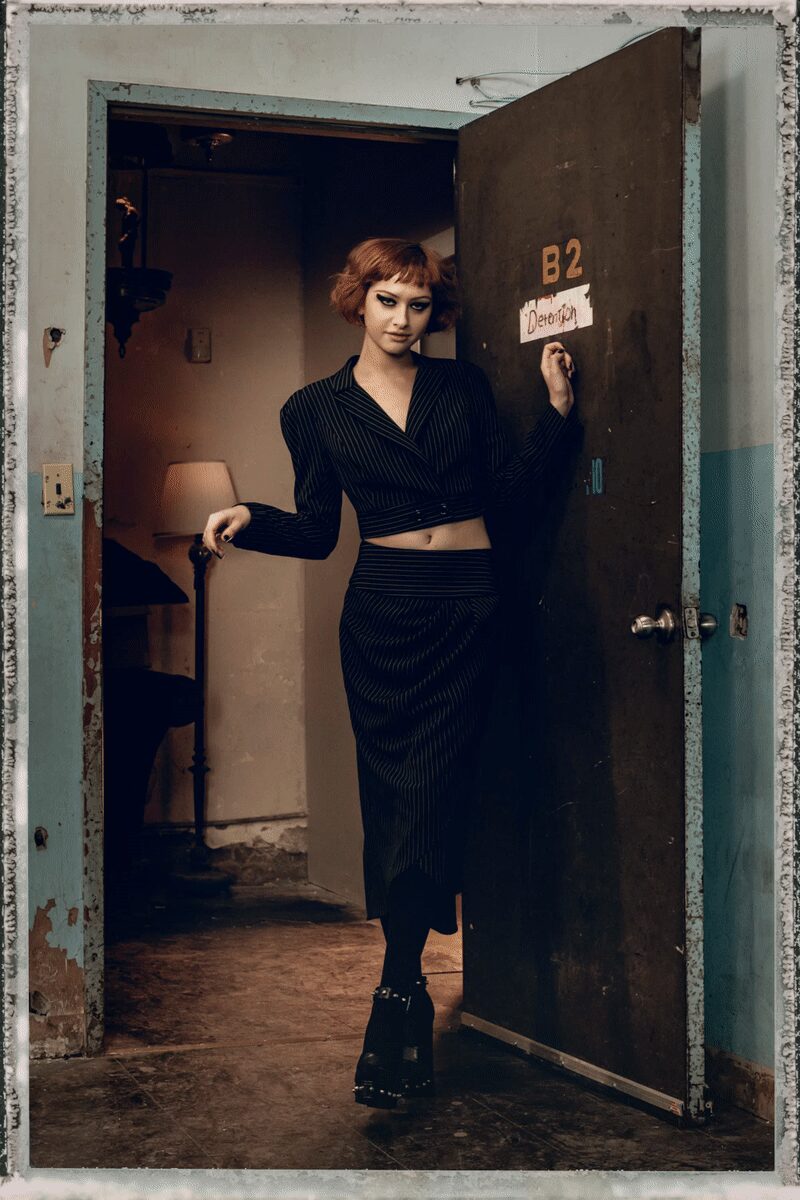 Black pinstriped cropped jacket, wrap front closing low across the ribs. Matching pinstriped pencil skirt with a broad waistband and uneven hem. Opaque black tights. Heavy black platforms with an ankle strap and metal studs. Answering the motel door at noon when checkout was hours ago.
