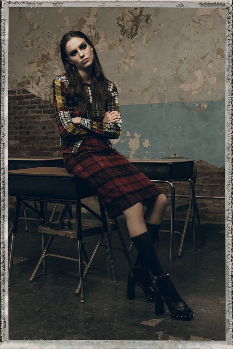 Patchwork plaid cardigan in mixed tartans, white placket running straight down center. Burgundy mohair plaid pencil skirt with a softly fuzzy surface. Black mid-calf socks. Black leather platform sandals, silver studs circling the sole. After-hours in an old classroom, radiators ticking against the cold.