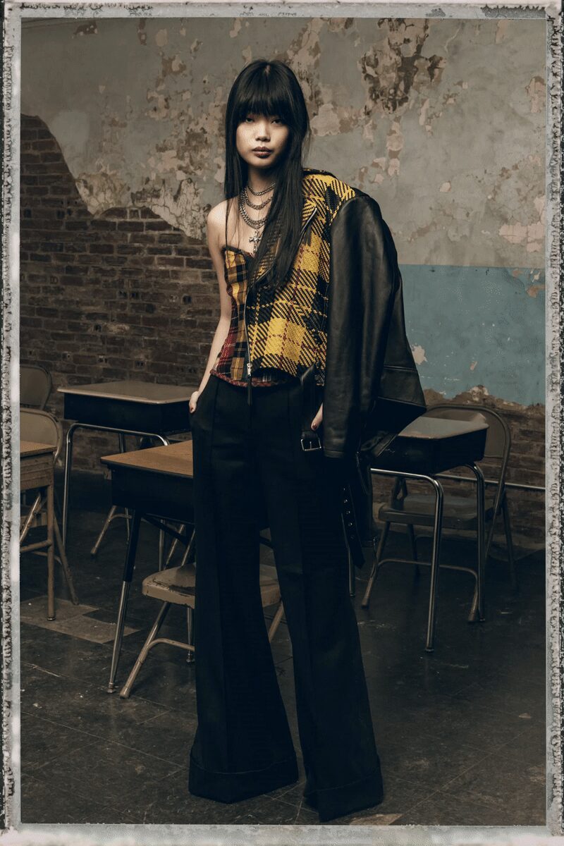 Asymmetric plaid corset top, one side cut away to the bare shoulder. Black leather jacket shrugged off one shoulder, thick belt hanging loose. Black wide-leg trousers with deep pleats and heavy turned-up cuffs. Layered silver chains and a small cross. After-hours classroom, chalk dust still floating above the desks.