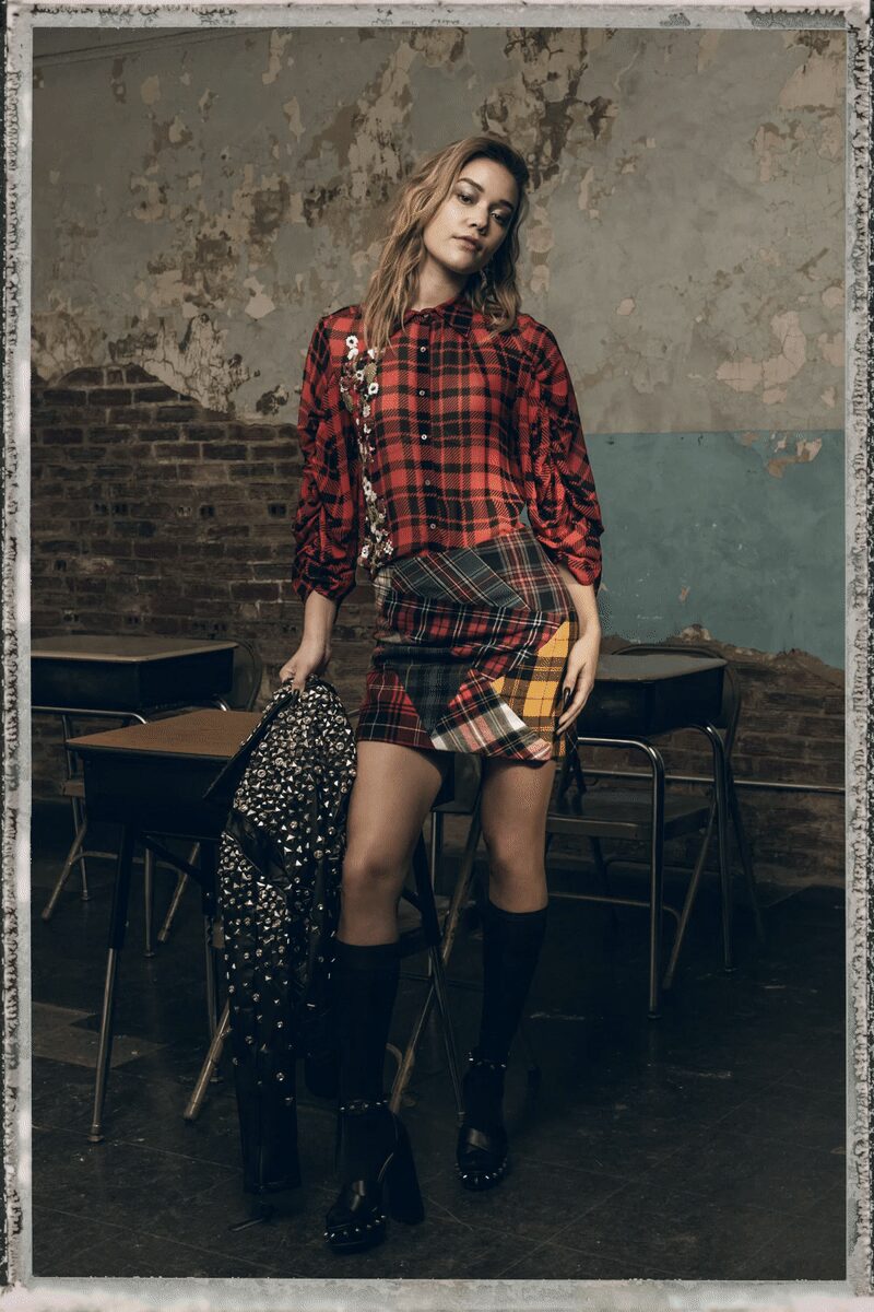 Red tartan shirt with a vertical spill of silver sequins. Patchwork plaid mini, panels crossing crooked at the hip. Black knee socks. Chunky black platforms ringed with tiny silver studs. Black leather jacket densely pressed with cone studs. After-hours in an abandoned classroom, desks shoved back against the wall.