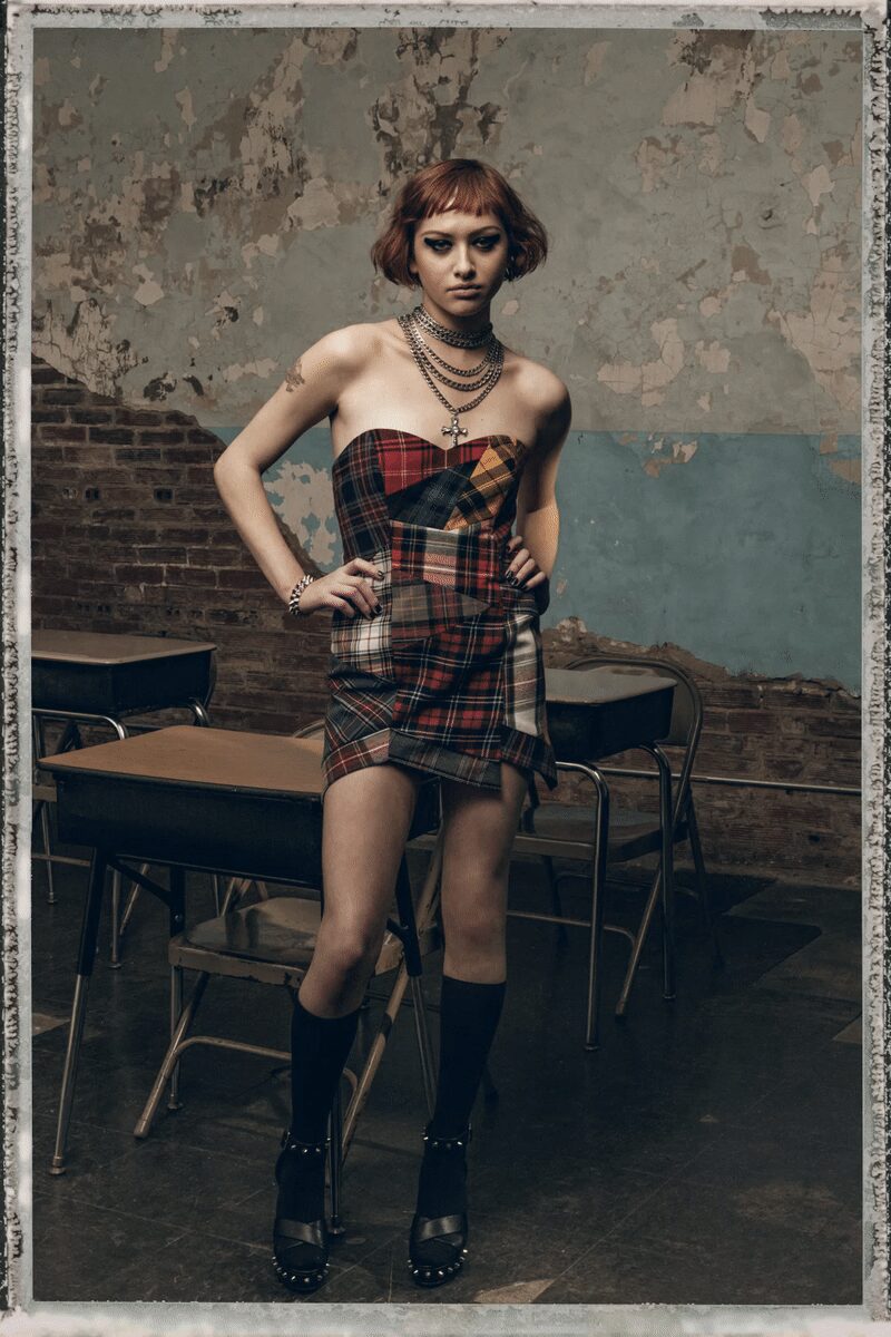 Patchwork tartan mini, folded on the bias with an uneven wrap hem. Stacked gunmetal chains with a dangling cross at the sternum. Chunky chain bracelet. Plain black knee socks. Black leather platforms ringed with silver dome studs. After detention, desks still warm and the radiator hissing.