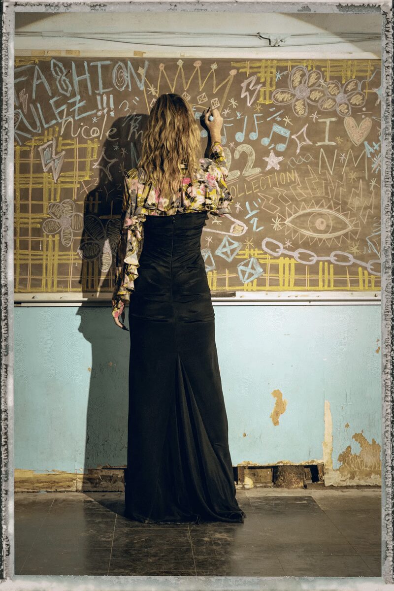 Black satin column gown, zip running straight down the bare back. Floral chiffon blouse worn off the shoulders, one sleeve ending in a ruffled cascade. A stack of mixed bangles at the raised wrist. Writing on a classroom chalkboard after everyone else has gone home.