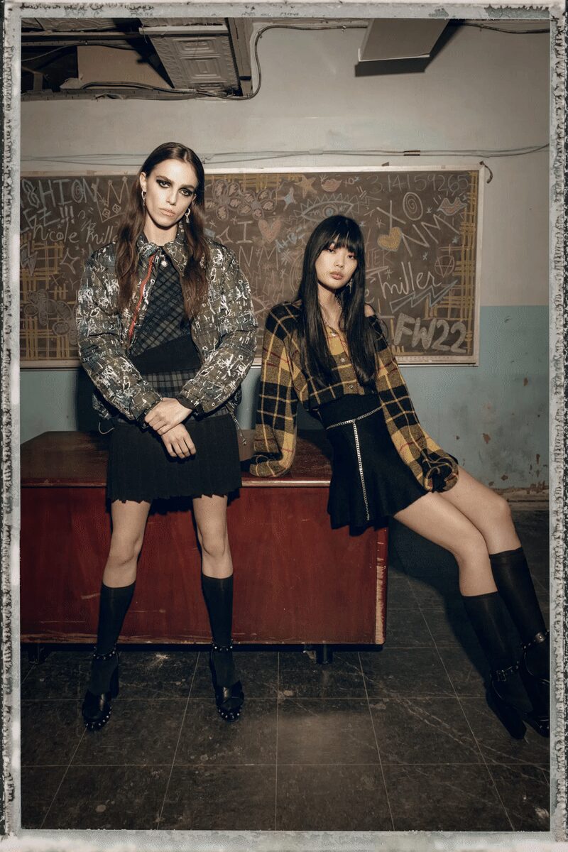 Grey bomber scribbled all over, red zip tape flashing at the placket. Black diamond-check knit top under a pointed collar. Black pleated mini with a raw, uneven hemline. Plain black knee socks. Chunky black loafers ringed with silver studs at the welt. Detention ended an hour ago, nobody has wiped the chalkboard clean.