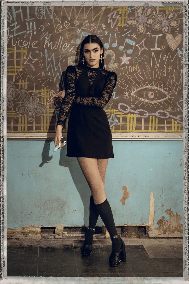 Black mini dress with deep V bib and plain A-line skirt. Sheer black lace underlayer, high neck and ruched sleeves. Long crystal cross earrings. Opaque black knee socks. Studded straps and heavy block heels. Leaning by the classroom blackboard after everyone else has gone home.