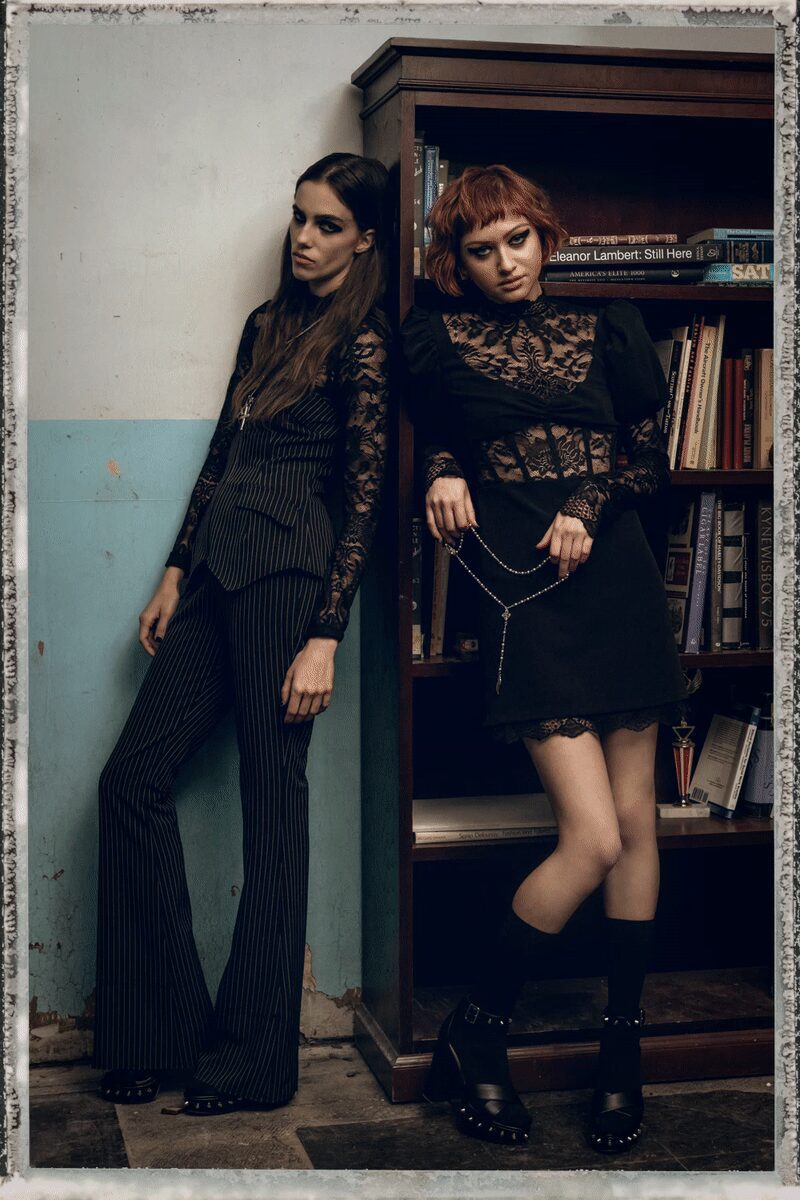 Black floral lace top, sheer through the sleeves and high neck. Charcoal pinstriped waistcoat, sharply pointed hem skimming the hips. Matching pinstriped flares breaking long over the platform shoes. Black platforms with a row of silver studs at the welt. Leaning in the library after the party has turned argumentative.