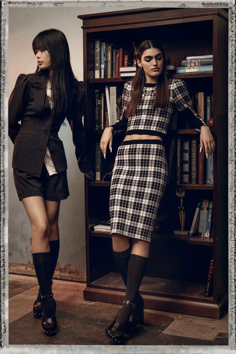 Black-and-ivory plaid knit crop top with sheer black sleeves. Layered silver chains and a long cross pendant. Matching plaid pencil skirt, hem landing below the knee. Dark knee socks. Black platform sandals with metal studs round the sole. Leaning on the den bookshelf after everyone else has chosen a game.