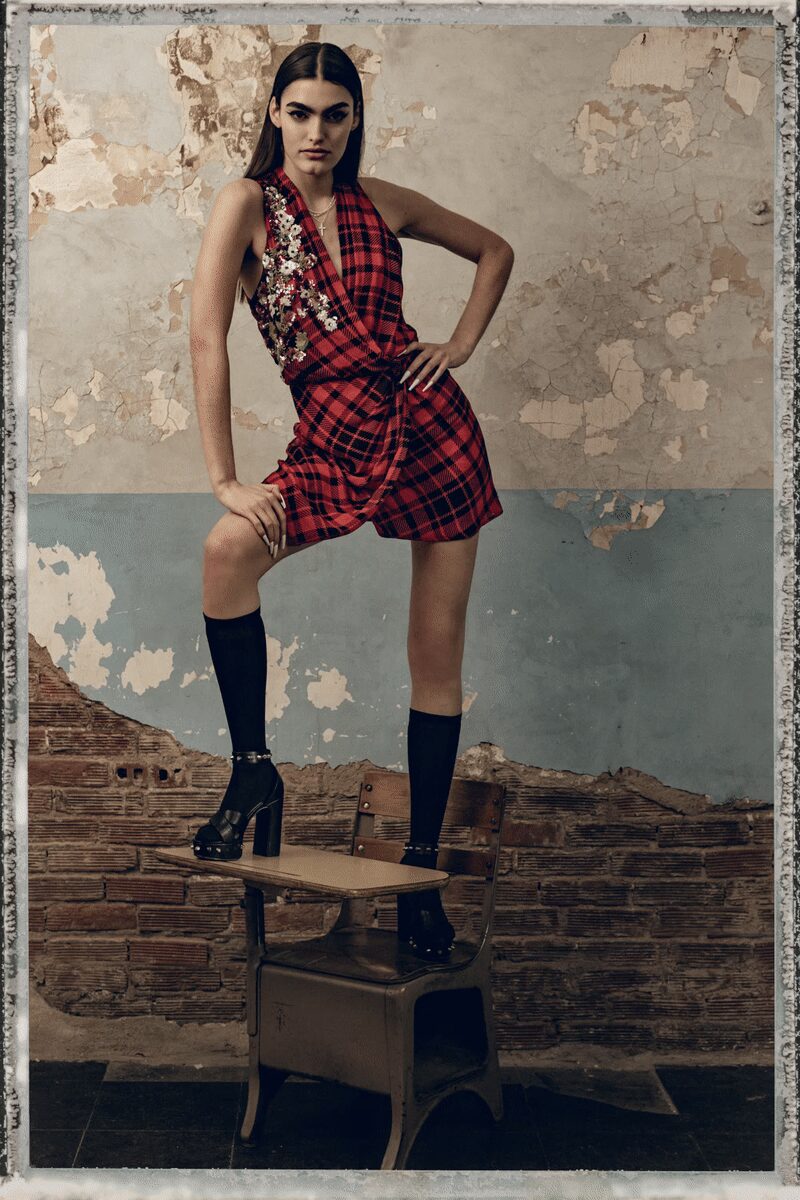 Red tartan wrap mini, one side crusted with silver floral paillettes. Fine chain cross necklace dropped into the plunging V. Black knee socks pulled straight and flat. Black leather platforms with round studs circling the ankle strap. Standing on a school chair after midnight when the party spills into the classroom.