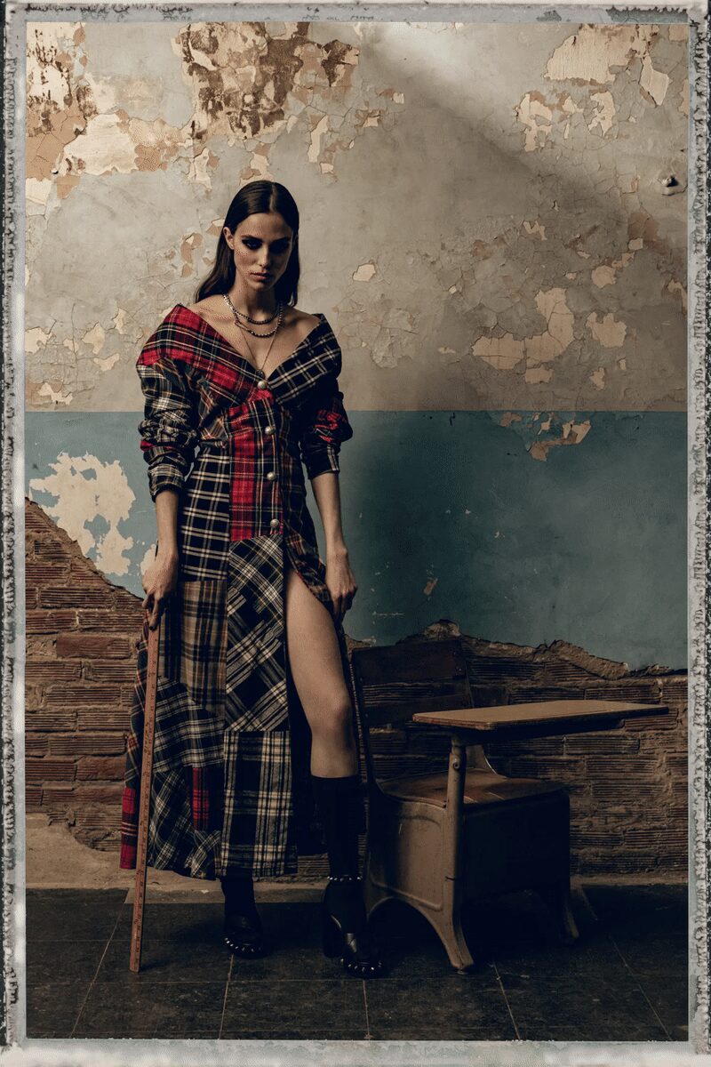 Off-shoulder patchwork plaid shirtdress, pearl buttons marching down a thigh-high slit. Layered gunmetal chains with one long drop between the collarbones. Black mid-calf platform boots ringed with small silver studs at the sole. After-hours in a disused classroom, plaster dust still lifting off the walls.
