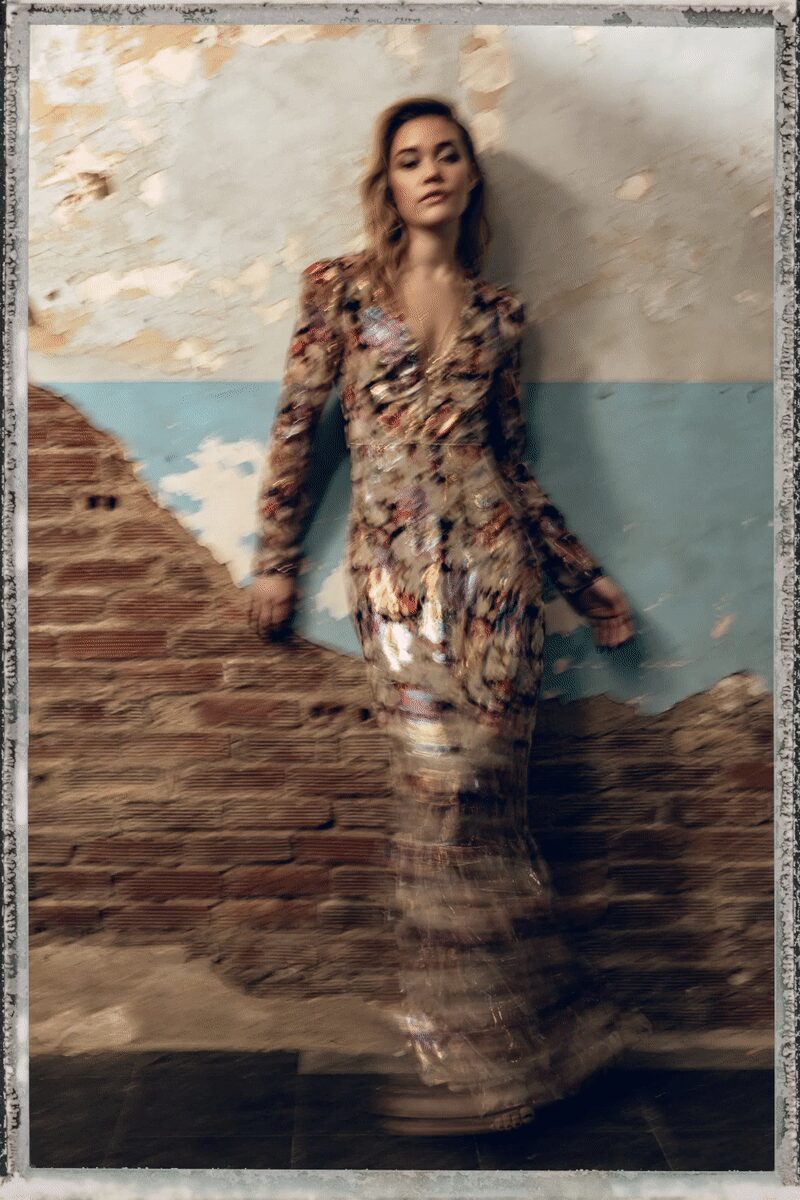 Long-sleeved plunging gown in mottled metallic paillettes, fluted into a floor-brushing hem. Barely-there platform sandals, straps ghosting at the blurred ankle. In a half-ruined seaside hotel after the band has packed up.