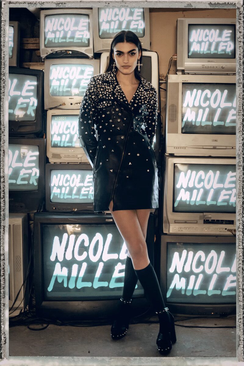 Black leather coatdress, collar and shoulders crusted with mixed silver studs. Long cross earrings grazing the jaw. Black ribbed knee socks. Thick platform Mary Janes ringed with dome studs at the sole. Waiting for the repair shop owner to admit none of these televisions work.