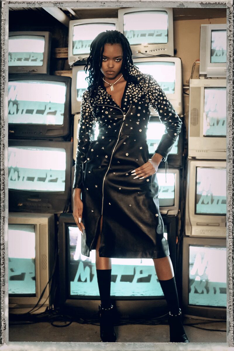 Black leather biker coat dress, shoulders and lapels crowded with silver cone studs. Short pearl strand with a dangling crystal cross. Single nude fishnet glove on one hand. Black knee socks. Glossy black platforms with a ring of metal studs at the ankle. Waiting for the arcade to open, CRT static already humming.