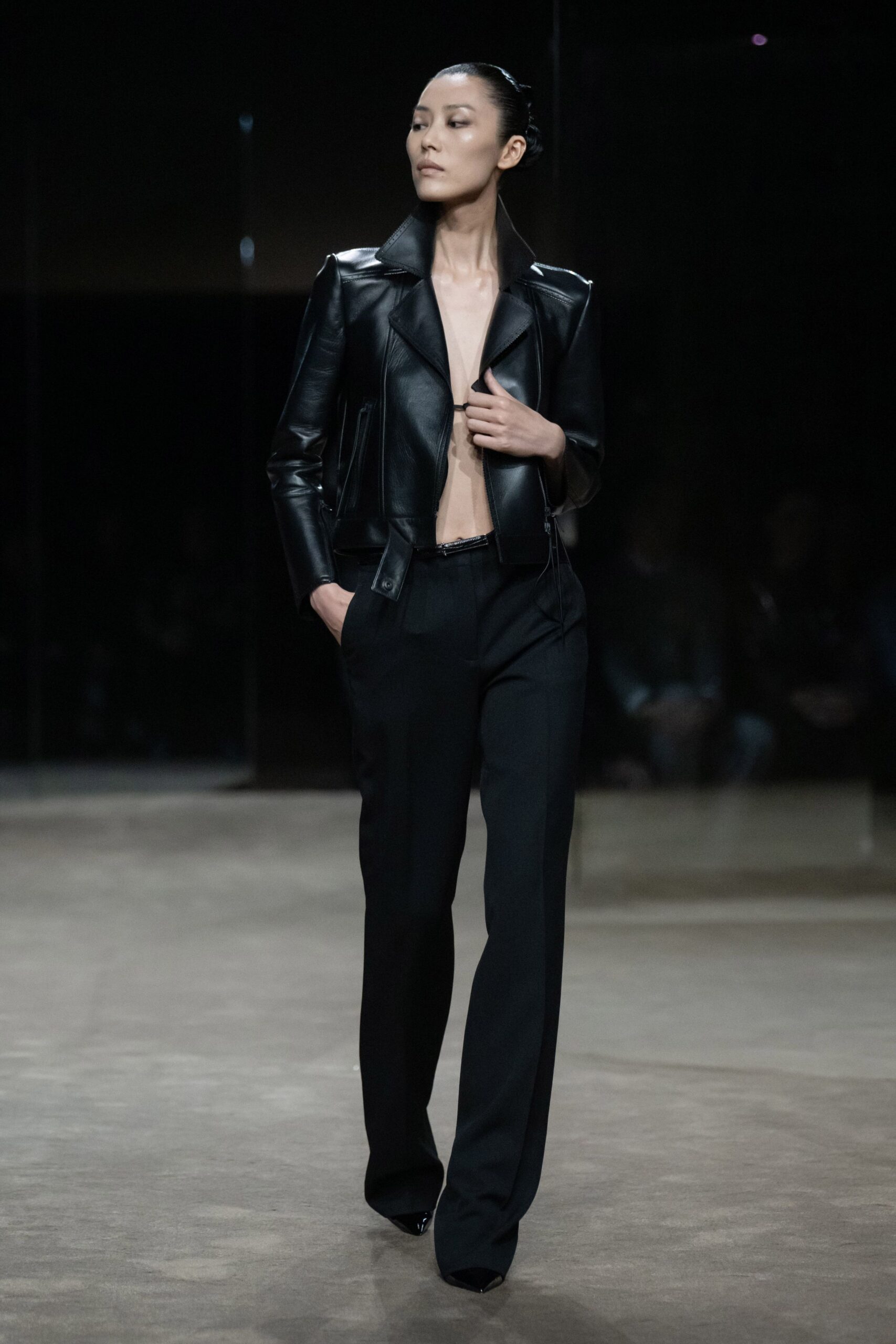 Black leather jacket cropped hard, with a high collar and long zip pulls. Black tailored trousers falling straight from a narrow waistband. Glossy pointed pumps, mostly swallowed by the hem. For walking into the hotel bar after midnight with nowhere else to be.