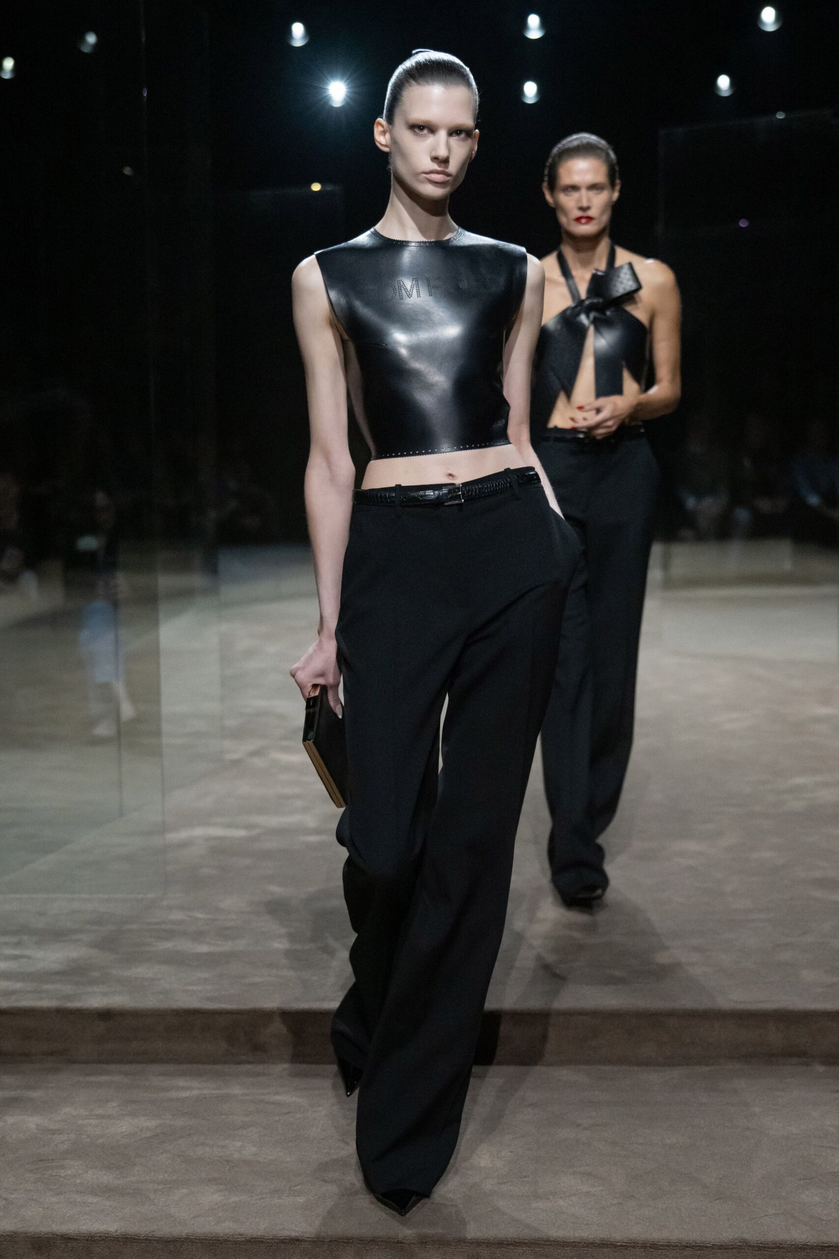 Black leather shell top, cropped hard at the waist with tiny perforated lettering. Low-slung black trousers falling loose from a narrow braided leather belt. Slim box clutch with a pale wood edge. Black pointed boots disappearing under the hem. Crossing a hotel lobby after midnight, still carrying the invitation.