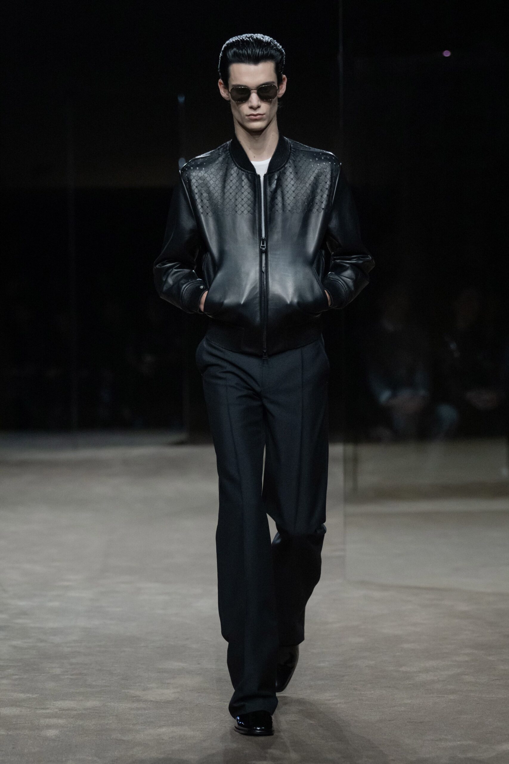 Dark oval lenses in a slim metal frame. Black leather bomber, woven paneling across the yoke and shoulders. White crewneck just visible at the collar. Ink-dark tailored trousers with a sharp front crease. Glossy black square-toe shoes. Walking through an empty office tower lobby after everyone else has gone home.