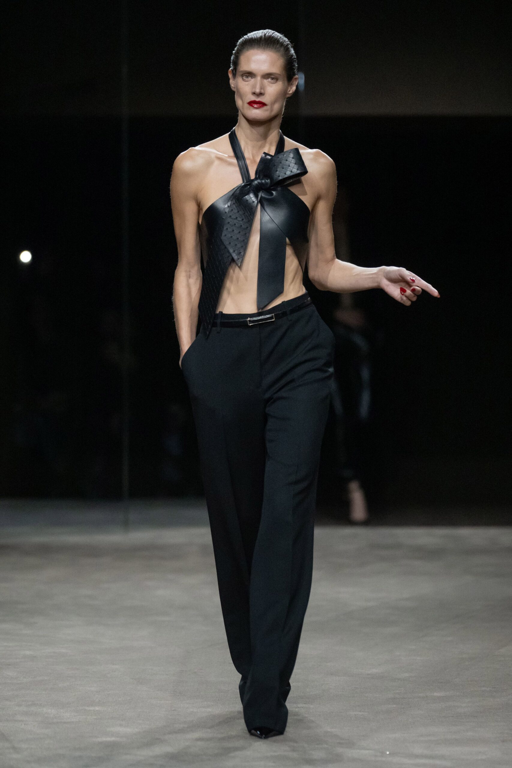 Black leather halter tied into an oversized knot, one panel basket-woven. Black tailored trousers, straight through the leg with handsunk pockets. Narrow belt with a slim rectangular silver clasp. Glossy black pointed pumps peeking under the hem. Whatever you wear home after missing the hotel bar's last call.