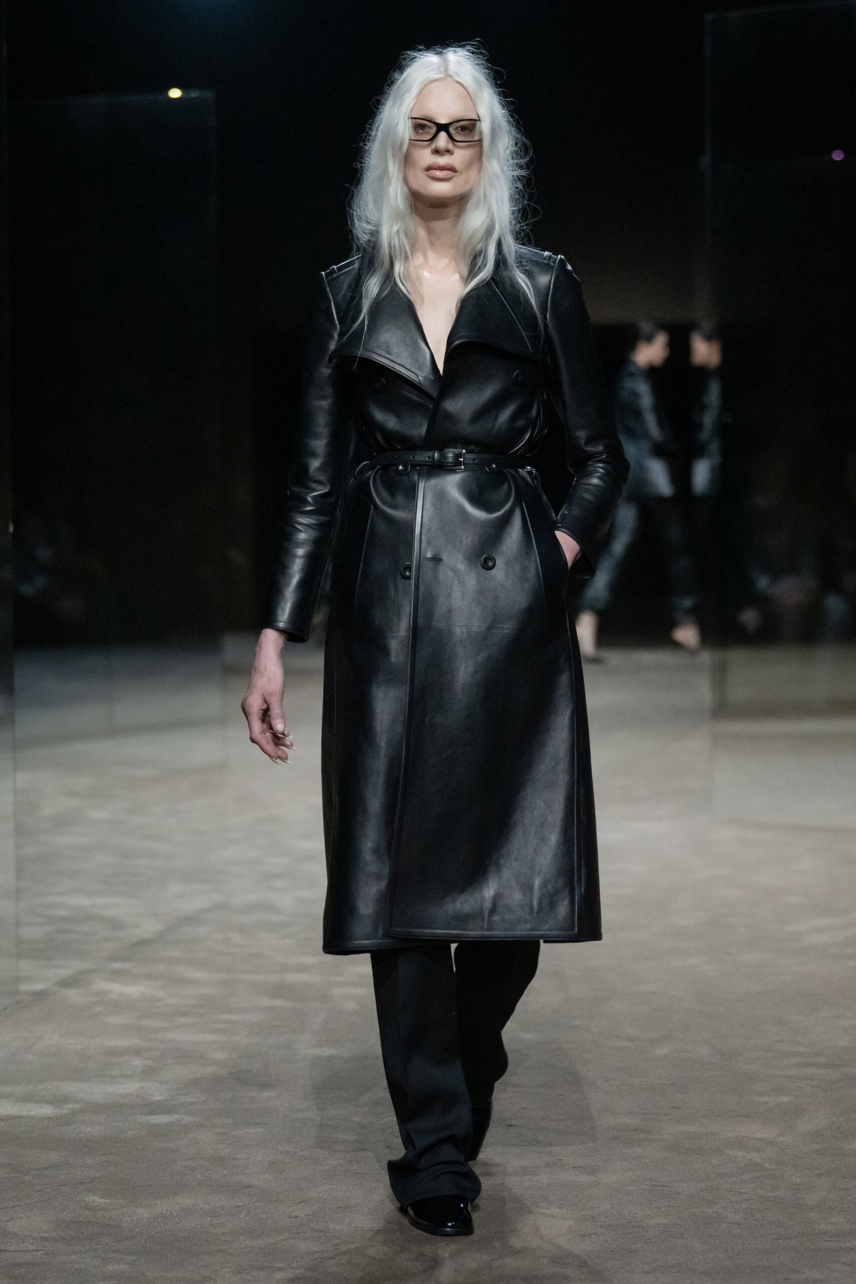 Rectangular black frames with a lightly smoked lens. Black leather trench dress, wide lapels and a skinny buckled waist belt. Black trousers breaking softly over the shoe. Glossy black square toes peeking from the hem. For signing papers in a lobby where every surface throws back your reflection.