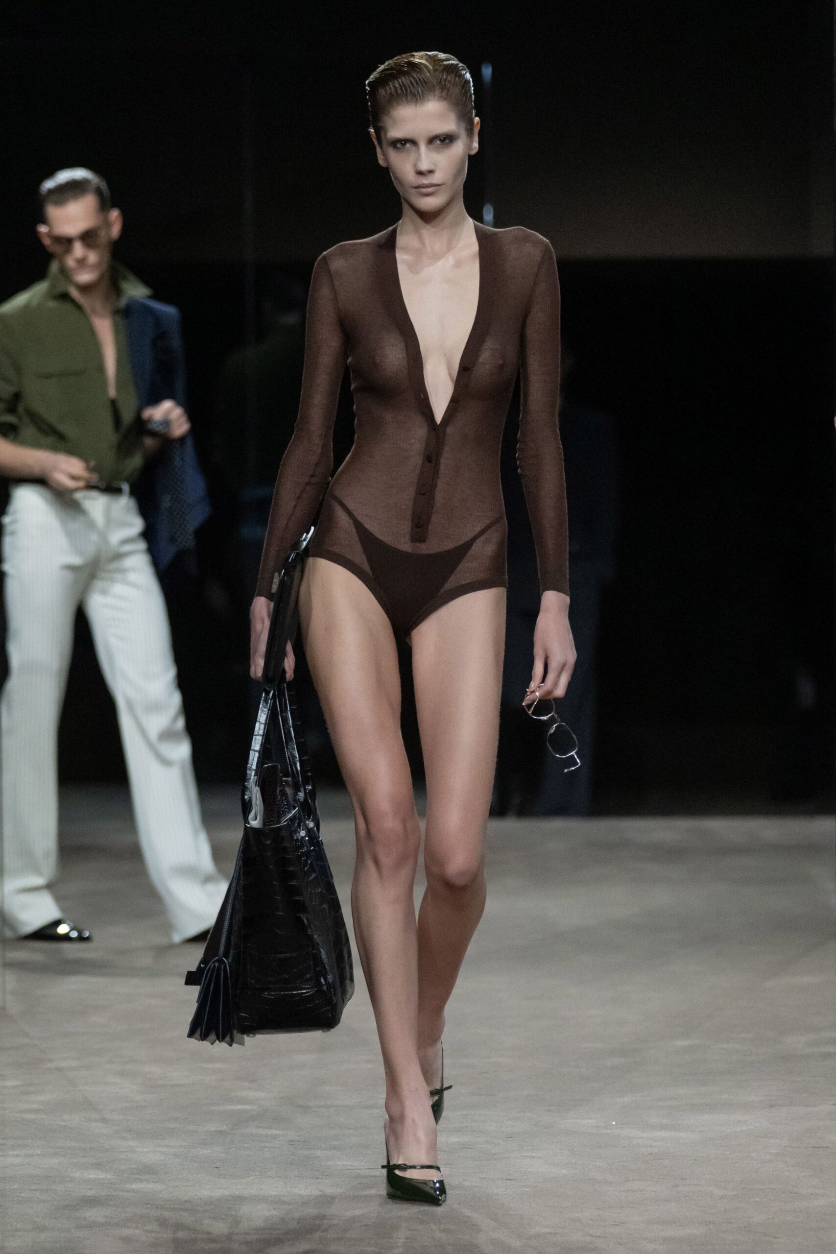 Sheer espresso knit bodysuit with a plunging buttoned V and darker brief panels. Black crocodile-embossed tote, accordion sides opening by the knee. Thin wire spectacles carried in hand. Black patent slingback pumps with a narrow strap. Walking back from the hotel pool after deciding not to get dressed.