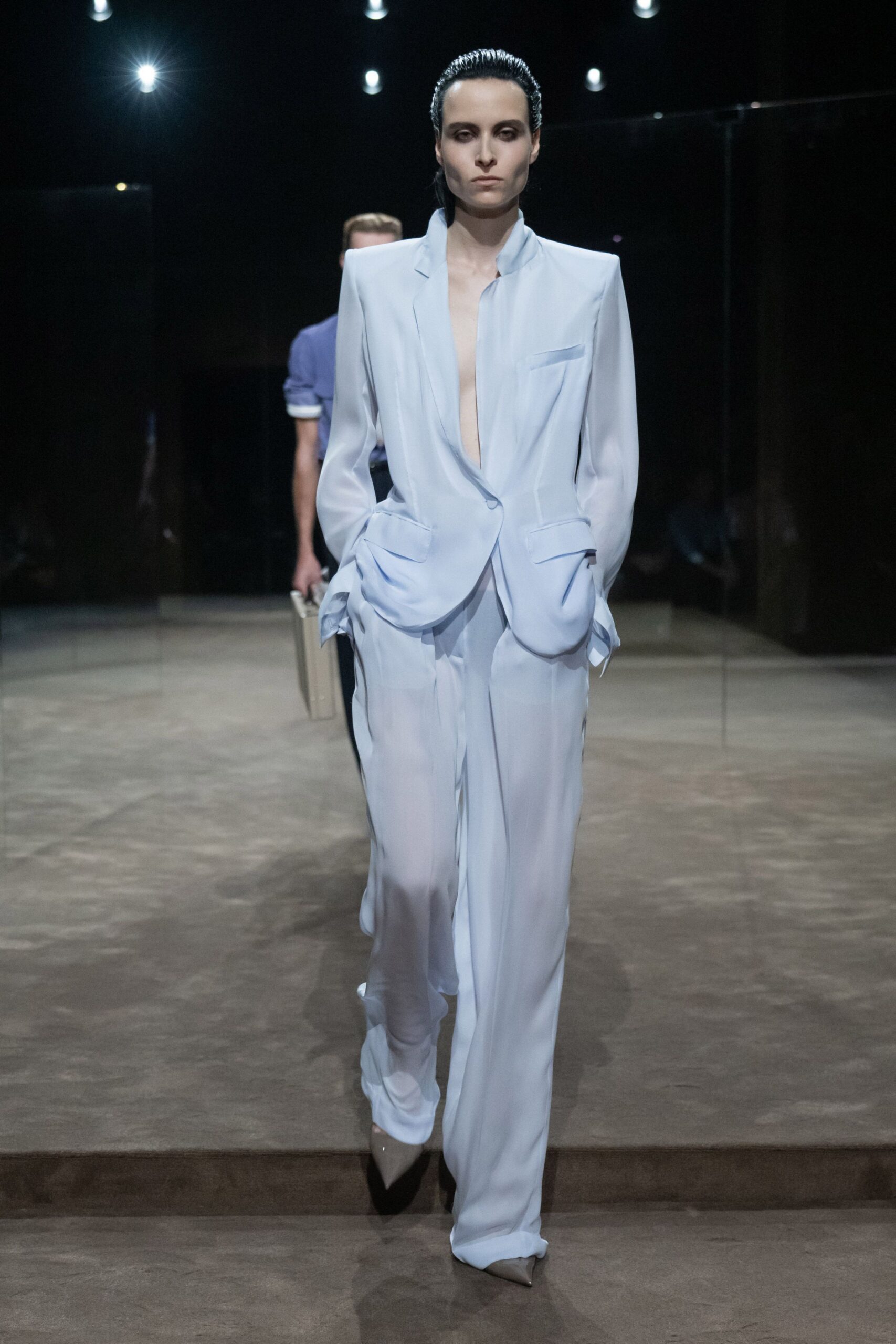 Powder-blue crepe blazer with a plunging front and skewed flap pockets. Matching fluid trousers breaking long over the shoe. Taupe pointed pumps, barely showing under the hem. For crossing a hotel lobby before sunrise with yesterday still on you.
