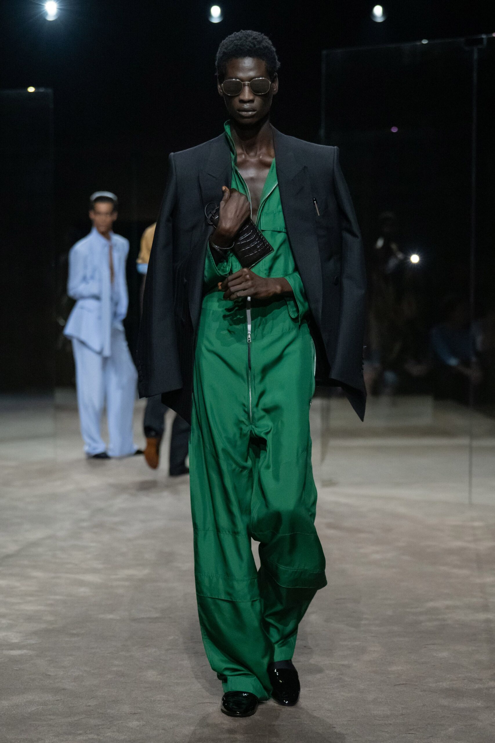Round smoke lenses in thin metal rims. Charcoal tailoring worn cape-like off the shoulders, a pen at the chest. Emerald zip-front jumpsuit with a deep split neckline and dropped rise. Black crocodile-embossed pouch looped at the wrist. Black patent shoes. Walking across an empty convention hall before the booths open.