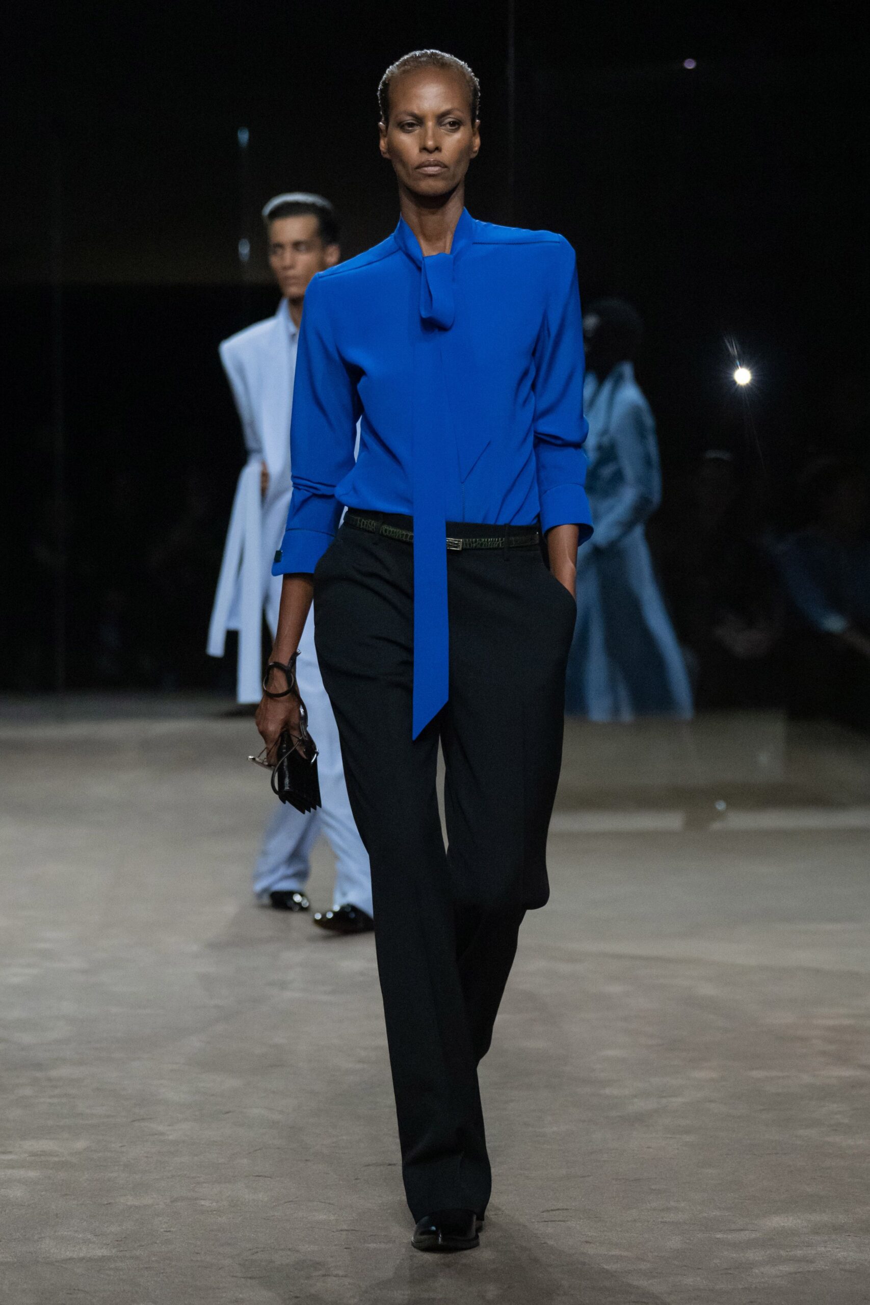 Electric blue blouse with a long scarf tie dropping past the waist. Black tailored trousers with a low belt and hands-deep pockets. Slim black belt with a small rectangular buckle. Tiny black wallet on a wrist loop. Black square-toe shoes. For the morning you have to look composed before speaking first.