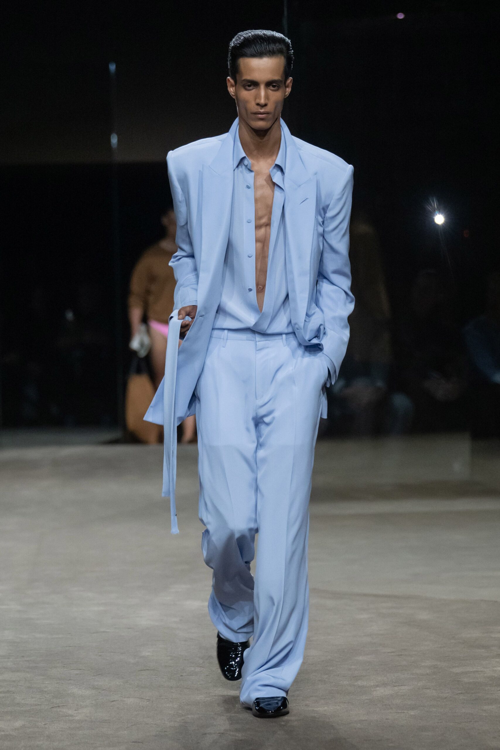 Powder-blue blazer with razor shoulders and a long sash slipping from the cuff. Matching fluid shirt, unbuttoned to the waist with pale snap buttons. Loose tailored trousers pooling softly over the shoes. Black patent square toes. Walking into a hotel lobby just after the wedding let out.